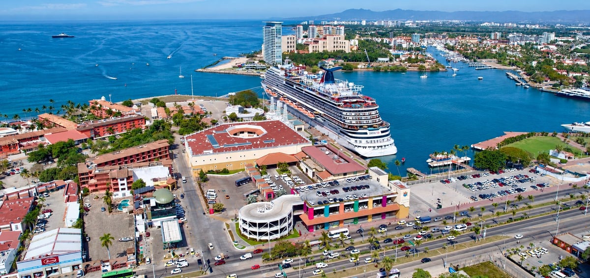 Aerial view of a cruise ship docked at a busy maritime terminal, surrounded by colorful buildings, parking areas and blue water under a clear sky
